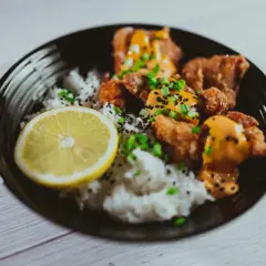 Chrupiący kurczak karaage z ryżem, polany sosem, posypany szczypiorkiem i czarnym sezamem, z plasterkiem cytryny.