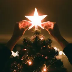 Hände setzen einen leuchtenden Stern auf die Spitze eines Weihnachtsbaums, der die weihnachtliche christliche Bedeutung des Lichts und der Hoffnung symbolisiert.
