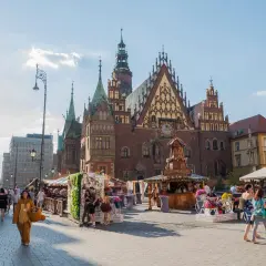 Tętniący życiem wrocławski Rynek, z Ratuszem w tle. Ludzie spacerują wśród kramów, a ktoś w stroju misia dodaje uroku temu miejscu.