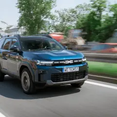 Niebieski Dacia Jogger na drodze, z widocznymi budynkami w tle. Marka samochodu na m.