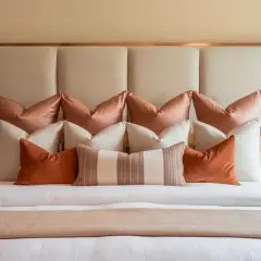 A masterclass in how to layer pillows on a bed, featuring a plush headboard and a symphony of rust, cream, and blush tones.