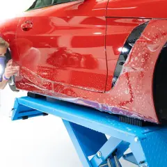 Man applicerar skyddsfilm på en röd bil under maskinpolering. Bilen lyfts upp på en blå billyft.