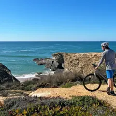 A cyclist pauses on a cliffside path during a Porto to Lisbon bike tour, admiring the vast blue ocean.