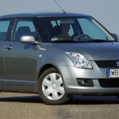 Szary Suzuki Swift 2007 z kierowcą za kierownicą, zaparkowany na drodze.