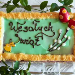 Zielone ciasto wielkanocne z napisem "Wesołych Świąt", ozdobione zajączkiem, baziami i pisankami.
