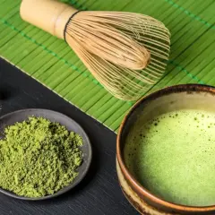 Matcha-Pulver auf einem Teller, daneben eine Schale mit schaumiger Matcha-Creme und ein Bambusbesen.