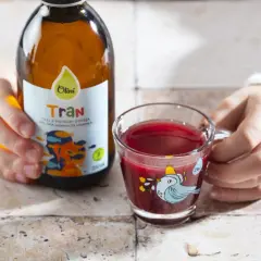 Dziecko trzyma butelkę z tranem dla dzieci Olini i szklankę z czerwonym napojem.