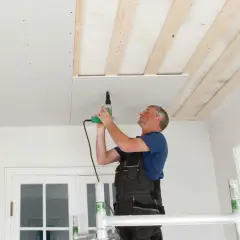 Man på byggställning skruvar gipsskivor i taket. Taket är under renovering med synliga träreglar.