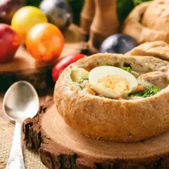 Pyszny barszcz biały lub żurek w chlebie, ozdobiony jajkiem. W tle kolorowe pisanki i bochenek chleba.