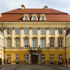 Elegancki budynek z żółtą fasadą i czerwonym dachem, ozdobiony licznymi oknami. To muzeum sztuki medalierskiej we Wrocławiu.