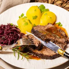 Zarter Rinderbraten im Ofen, serviert mit Salzkartoffeln und Rotkohl. Ein Messer und eine Gabel sind bereit, das saftige Fleisch anzuschneiden.