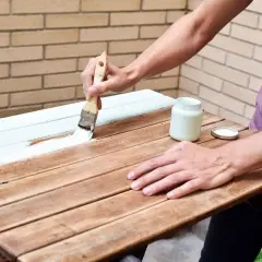 Persona pintando una mesa de madera exterior con brocha y pintura blanca.