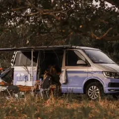 Un homme se détend près d'un camping-car sauvage, avec une table et des chaises installées à l'extérieur. Le véhicule est garé dans un cadre naturel verdoyant.