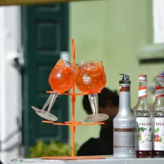 Dwa kieliszki z drinkiem Aperol Spritz na pomarańczowym stojaku, obok butelki syropów Monin.