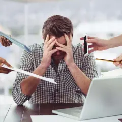 Pracownik przytłoczony zadaniami, z rękami na głowie, doświadcza napadów potu. Wokół niego krążą dokumenty, telefon i ołówek.