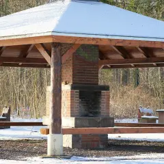 En träpaviljong med snö på taket och en murad grill i mitten. Bänkar och bord står redo för en mysig fika utomhus.