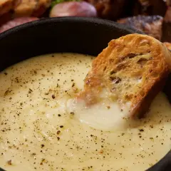 Una fetta di pane croccante si tuffa in una cremosa fonduta di formaggio, cosparsa di pepe nero. Sullo sfondo, salumi e pane tostato invitano a un convivio.