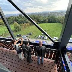 Widok z wieży widokowej na malowniczy krajobraz z lasami i polami. Na dole widać zaparkowane samochody, namioty i ludzi, jakby szykowali się na festyn lub zlot. Może to być nawet zlot motocyklowy, bo jeden z panów ma na sobie kamizelkę z napisem "GÓRY ...