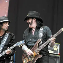 Lemmy Kilmister z zespołem na scenie. Dwóch muzyków gra na gitarach, jeden w kapeluszu, drugi w kowbojskim.