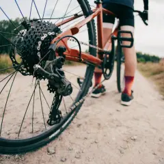 Widok z bliska tylnej przerzutki rowerowej, która wygląda na Shimano Acera lub Alivio, na tle szutrowej drogi i rowerzysty.