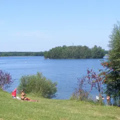 Słoneczny dzień nad jeziorem Chechło-Nakło. Kobieta odpoczywa na trawie, w tle błękitna woda i zielone wyspy.