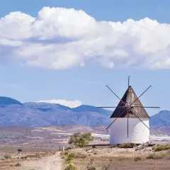 Molino blanco con aspas de madera bajo un cielo azul con nubes. Paisaje árido con montañas al fondo, como si Fernan Perez hubiera pintado este cuadro.