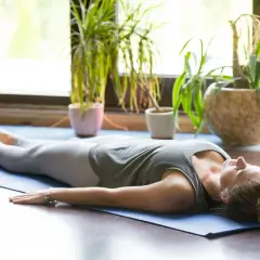 Kobieta w pozycji savasana, zrelaksowana na macie do jogi, otoczona zielenią roślin.