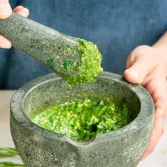 Frische Kräuter werden im Mörser zermahlen, um selbstgemachtes Pesto herzustellen. Ein Tropfen grüner Genuss fällt vom Stößel in die Schüssel.