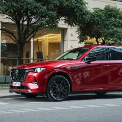 Lśniąca, czerwona Mazda CX-60 3.3 diesel na tle nowoczesnego budynku i drzew.