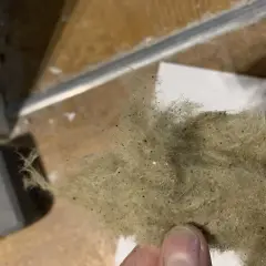 En hand håller en tuss av stenull eller glasull, som är dammig och innehåller små svarta prickar.