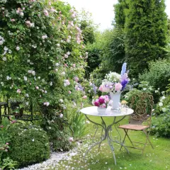 Ein romantischer Garten mit blühenden Rosen, ein Tisch mit Blumenarrangements und ein schmiedeeiserner Stuhl laden zum Verweilen ein. Ein idyllischer Ort, um einen **beet anlegen**-Traum zu verwirklichen.