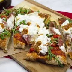 Pyszna pinsa z burrata, rukolą i szynką parmeńską, podana na desce.