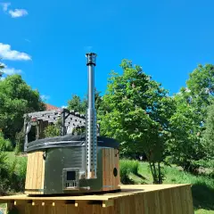 Drewniane jacuzzi w ziemi z metalowym kominem, otoczone zielenią i błękitnym niebem. Idealne miejsce na relaks.