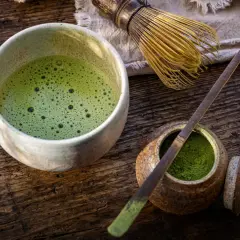 Un bol de matcha mousseux, une cuillère en bambou et de la poudre verte dans un pot sur une table en bois.