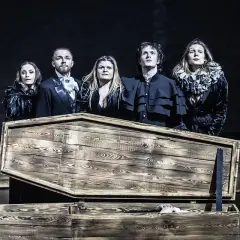Aktorzy Teatru Pantomimy Wrocław w strojach z epoki stoją nad otwartą trumną, z której wystaje ręka.