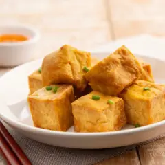 Tofu crujiente dorado, cortado en cubos y adornado con cebollín, servido en un plato blanco con salsa y palillos.