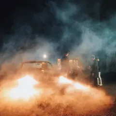 A film crew uses artificial light examples, including a bright spotlight and car headlights, to create a smoky, dramatic scene with men in hats gathered around a vintage car.