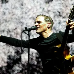 Bryan Adams na koncercie, śpiewa do mikrofonu i gra na gitarze.