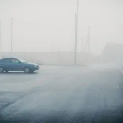 Un coche azul solitario se detiene en medio de una **niebla espesa** que oculta un camino y una casa.