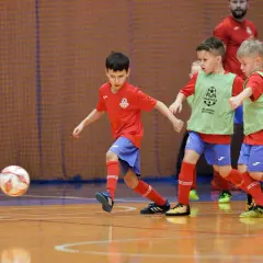 Zacięte współzawodnictwo sportowe dzieci i młodzieży na hali. Mali piłkarze w czerwonych koszulkach i zielonych kamizelkach walczą o piłkę.