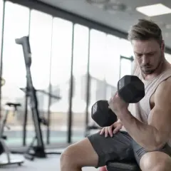Mężczyzna ćwiczy biceps z hantlem, pokazując mięśnie synergistyczne przykłady. W tle siłownia.