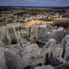 Ruiny zamku Ogrodzieniec górują nad okolicą, ukazując kamienne mury i łuki. W tle widać wioskę i lasy.