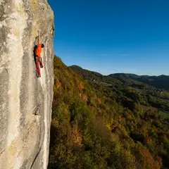 Ein Kletterer in orangefarbenem Shirt und roten Hosen erklimmt eine Felswand in Kroatien, während sich im Hintergrund ein herbstliches Tal erstreckt.