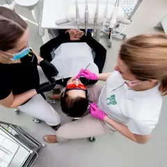 Dwie asystentki stomatologiczne pracują przy pacjencie. Jedna z nich, w czarnej bluzce, przygotowuje narzędzia. Druga, w białej koszulce, wykonuje zabieg.