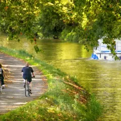 Pareja en bicicleta disfruta de un **canal du midi recorrido** bajo árboles frondosos, mientras una barcaza navega por el agua.