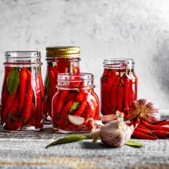 Słoiki z papryczkami chili w zalewie, obok czosnek i świeże papryczki. Idealne do pikantnych potraw.