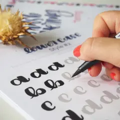 Mano practicando lettering con un rotulador negro sobre una hoja con letras "a", "b", "c" y "d" para calcar.