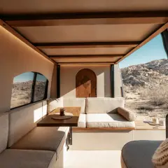 Intérieur de camping-car aménagé avec banquettes, table et porte en bois, vue sur un paysage désertique.