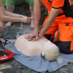 Ćwiczenie RKO na fantomie. Ratownik w pomarańczowym stroju uciska klatkę piersiową, a druga osoba przygotowuje AED.
