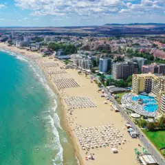 Słoneczna riwiera bułgarska z szeroką, piaszczystą plażą pełną leżaków i parasoli, turkusowym morzem i hotelami w tle.